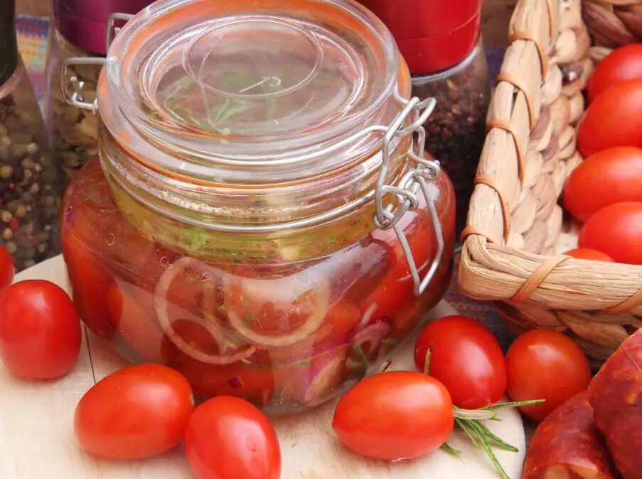 Ein Einmachglas mit eingelegten Tomaten, Zwiebeln und Gewürzen auf einem Brett, umgeben von frischen Tomaten und Wurst.