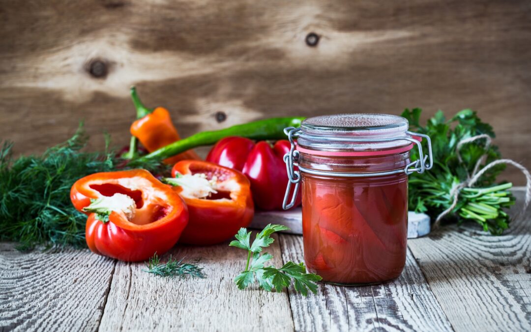 Verschlossenes Glas mit eingelegten Paprikaschoten, frische Paprika, Kräuter und Gemüse auf einem rustikalen Holztisch.