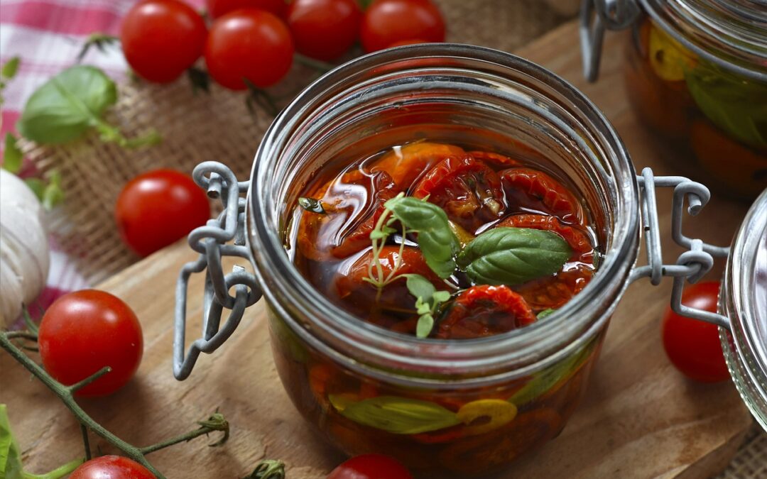 Ein Einmachglas mit getrockneten Tomaten und Basilikum, umgeben von frischen Kirschtomaten auf Holztisch.