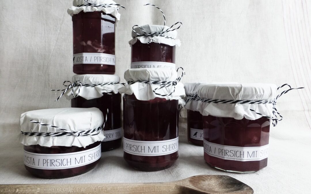 Sechs Marmeladengläser mit weißem Stoff bedeckt, gefüllt mit Josta-Pfirsich-Sherry-Mischung, davor ein Holzlöffel.