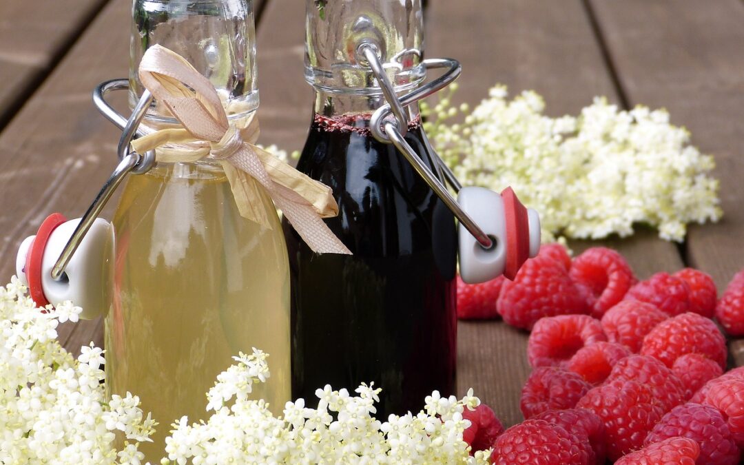 Zwei Flaschen Sirup, eine hell, eine dunkel, umgeben von frischen Himbeeren und Holunderblüten auf Holztisch.
