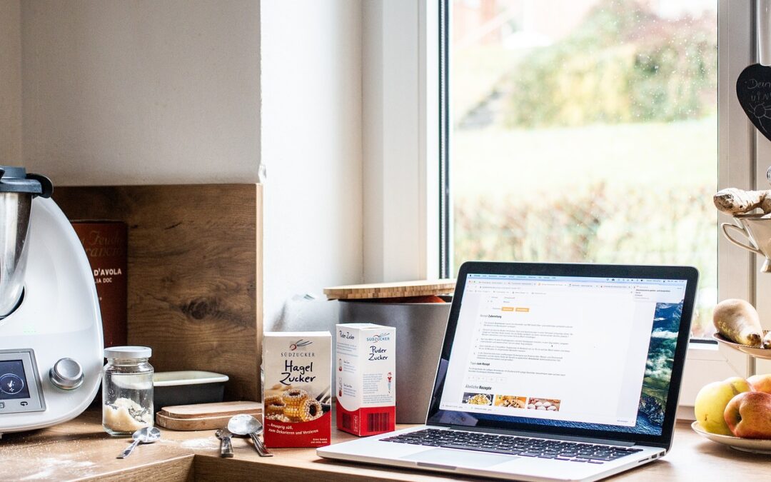 Laptop mit Rezeptanzeige neben Küchenmaschine, Zuckerpackungen und Äpfeln auf Holzarbeitsfläche vor Fenster.