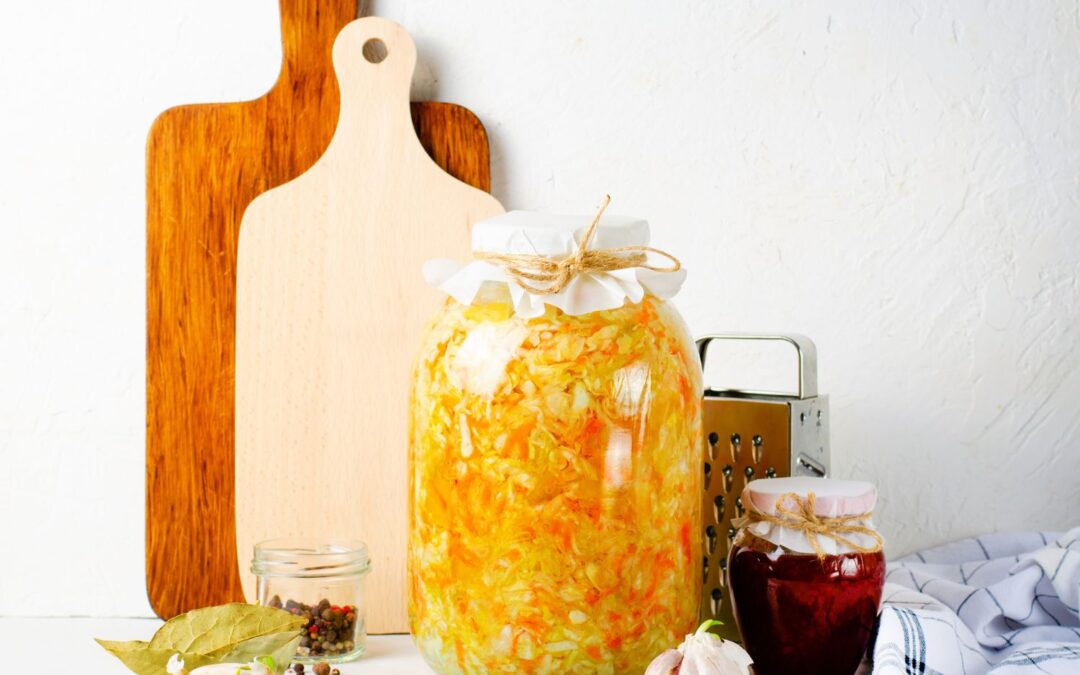 Glas mit eingelegtem Sauerkraut neben Gewürzen, zwei Schneidebrettern und einer Reibe auf weißem Untergrund.