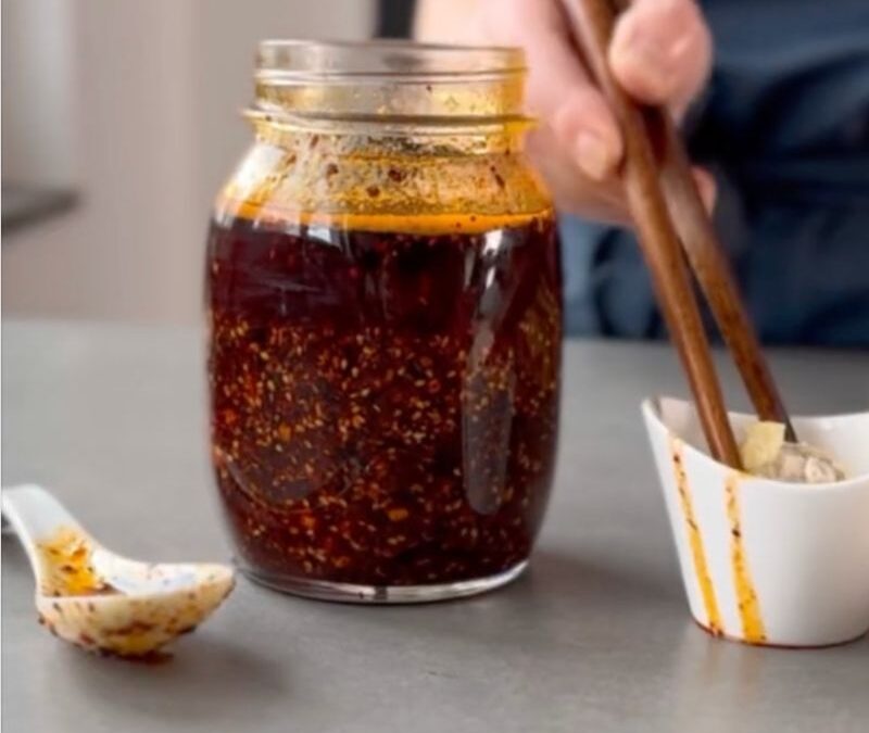 Ein Glas mit roter Chilisauce, eine Schale mit Essstäbchen und ein Löffel liegen auf einem grauen Tisch.