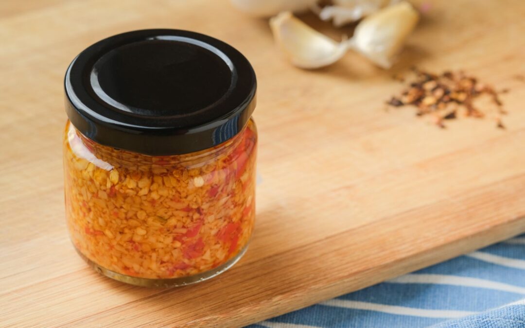 Ein Glas mit Chiliflocken und Öl auf einem Holzbrett, im Hintergrund Knoblauchzehen und Chiliflocken.