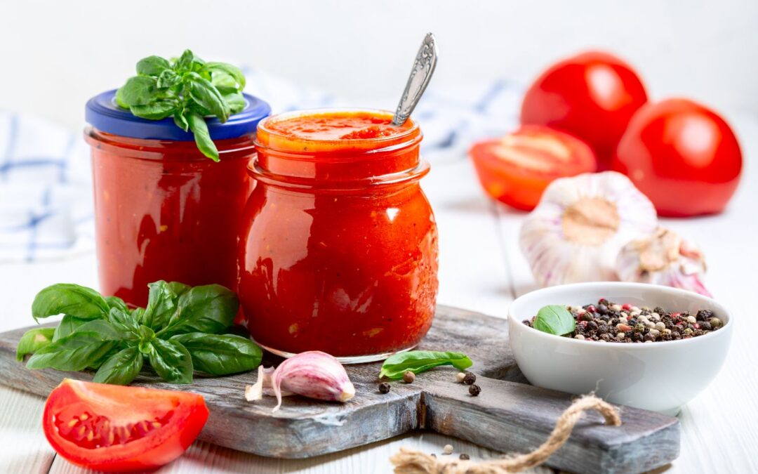 Zwei Einmachgläser mit Tomatensauce, frisches Basilikum, Knoblauch, Tomaten und Pfefferkörner auf einem Holzbrett.