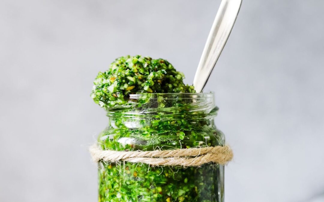 Glas mit grünem Pesto, Löffel steckt darin, mit Schnur umwickelt, vor grauem Hintergrund.