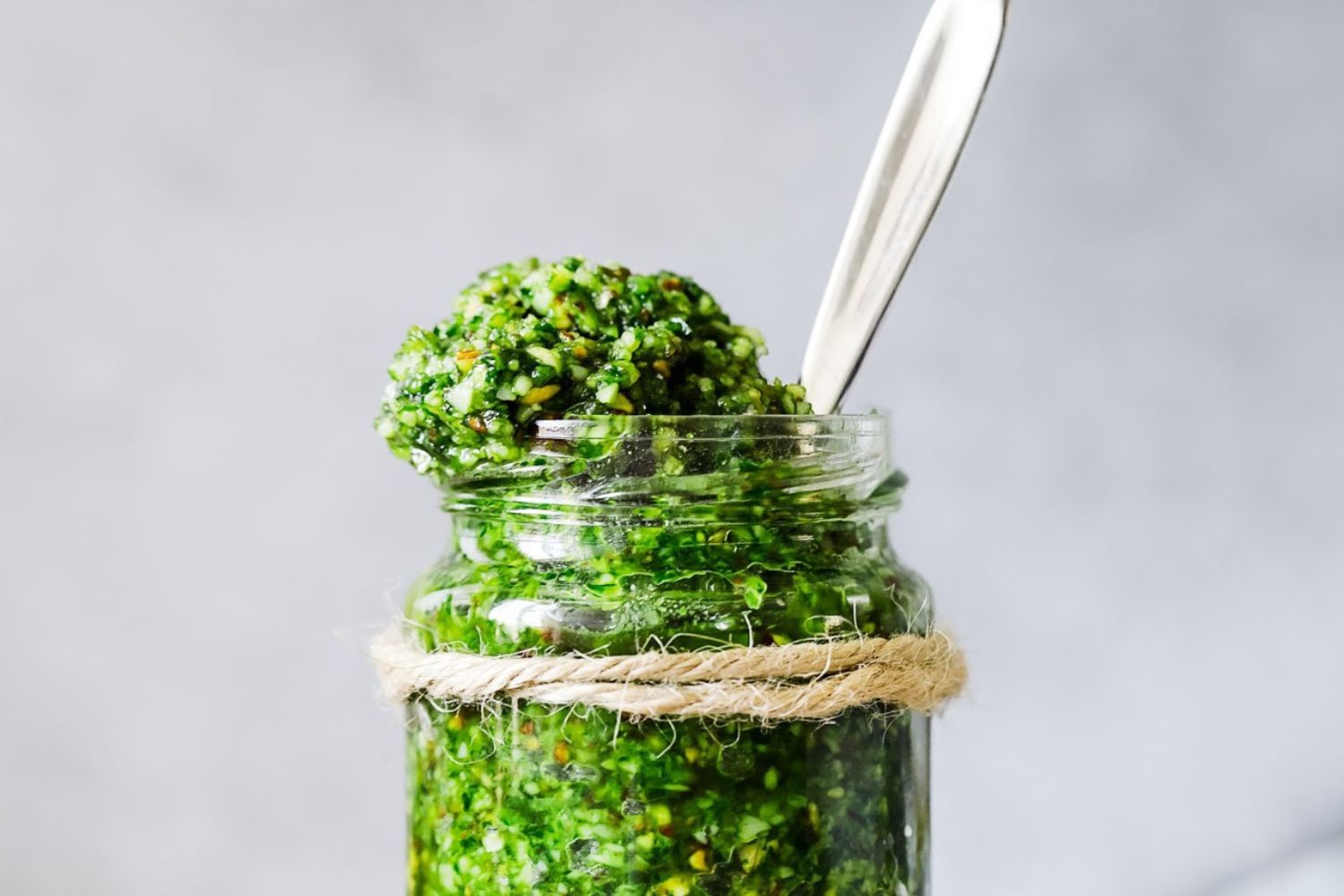 Bärlauch Pesto Glas mit grünem Pesto, Löffel steckt darin, mit Schnur umwickelt, vor grauem Hintergrund.