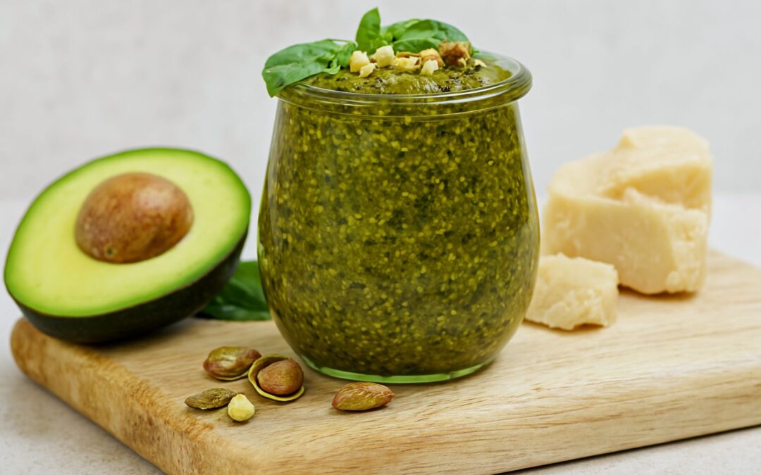 Ein Glas grünes Pesto mit Basilikum, Avocado, Käse und Nüssen auf einem Holzbrett arrangiert.