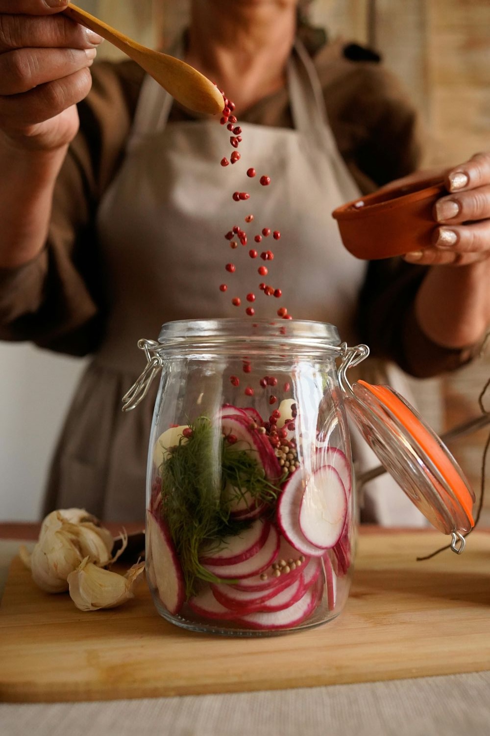 Radieschen fermentieren Person in Schürze gibt rote Beeren in ein Glas mit Radieschen, Kräutern und Knoblauch auf einem Holzbrett.