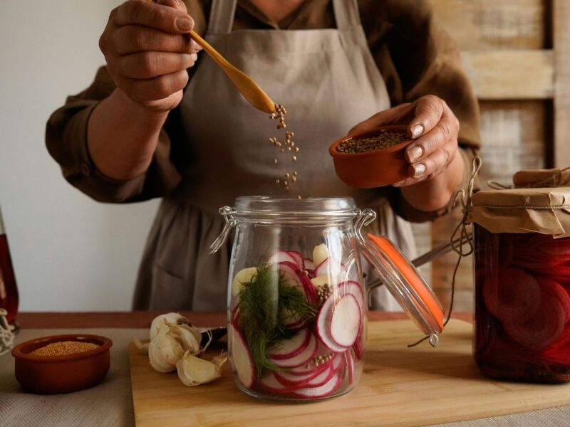Person füllt Gewürze in ein Glas mit Radieschen und Dill zum Einlegen. Daneben Knoblauch und ein weiteres Glas.