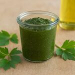 Glas mit grünem Pesto auf Holzuntergrund, dekoriert mit Petersilienblättern. Im Hintergrund eine Flasche Öl.