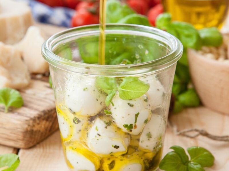 Mozzarellakugeln mit Kräutern in einem Glas, das mit Olivenöl übergossen wird. Im Hintergrund Tomaten und Basilikum.