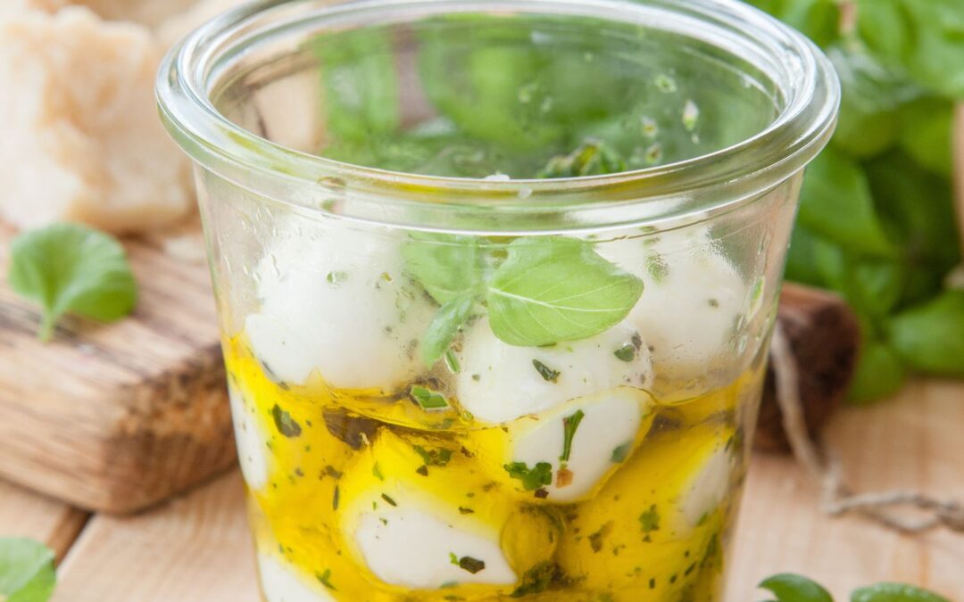 Glas mit eingelegten Mozzarellakugeln und Kräutern in Öl, im Hintergrund frisches Basilikum und Brot.
