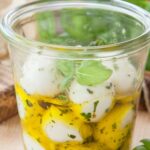 Glas mit eingelegten Mozzarellakugeln und Kräutern in Öl, im Hintergrund frisches Basilikum und Brot.
