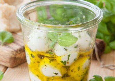 Glas mit eingelegten Mozzarellakugeln und Kräutern in Öl, im Hintergrund frisches Basilikum und Brot.