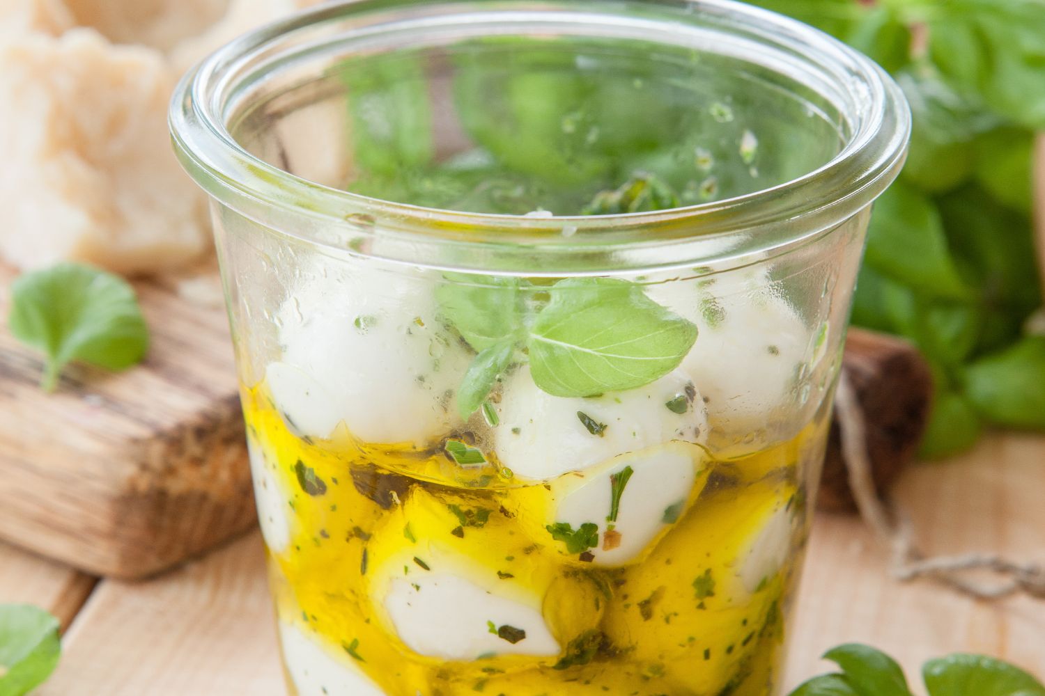 Eingelegte Mozzarella-Kugeln Glas mit eingelegten Mozzarellakugeln und Kräutern in Öl, im Hintergrund frisches Basilikum und Brot.