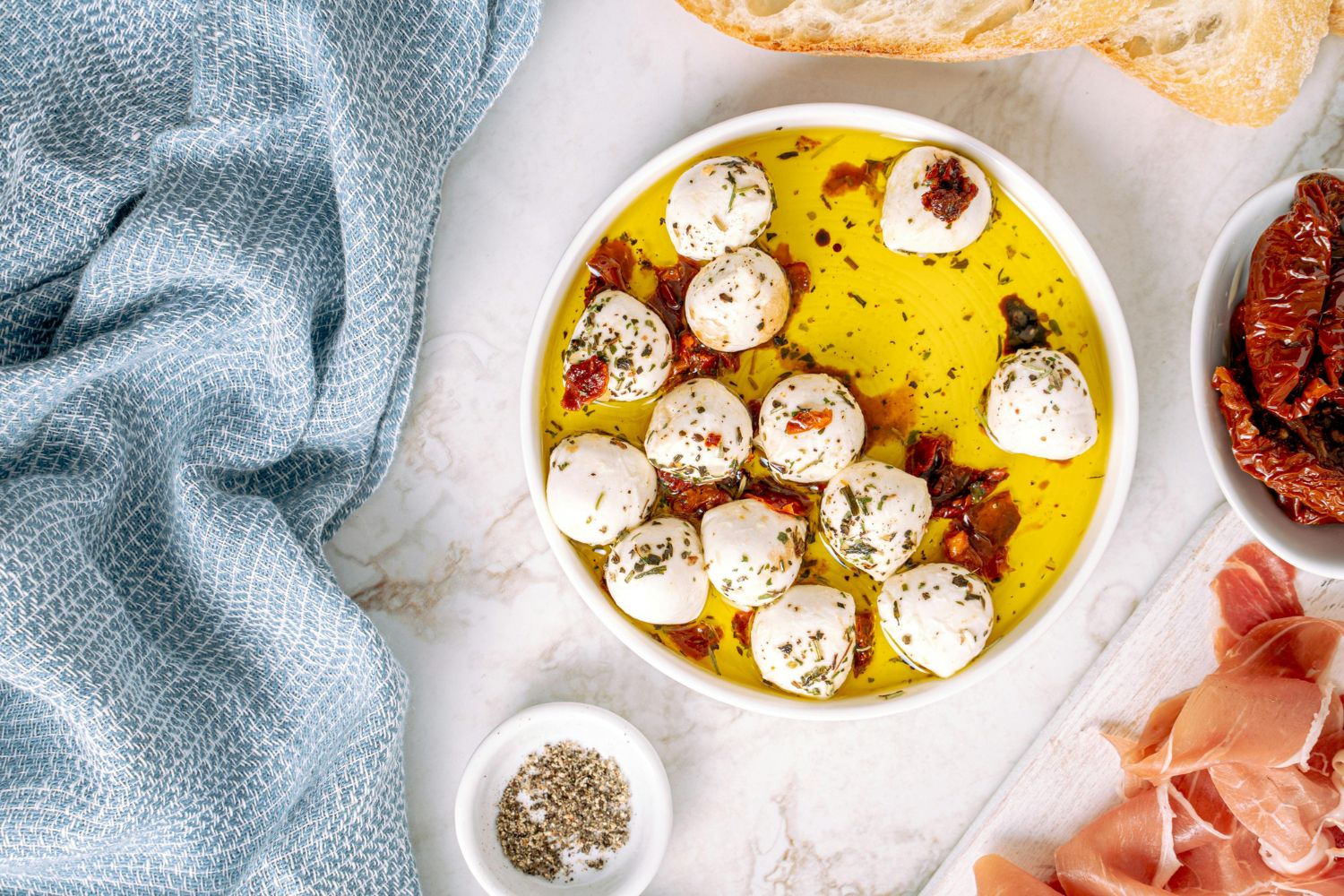 Kleine Mozzarellakugeln in Öl mit Kräutern und getrockneten Tomaten, umgeben von Schinken, Brot und Gewürzen.