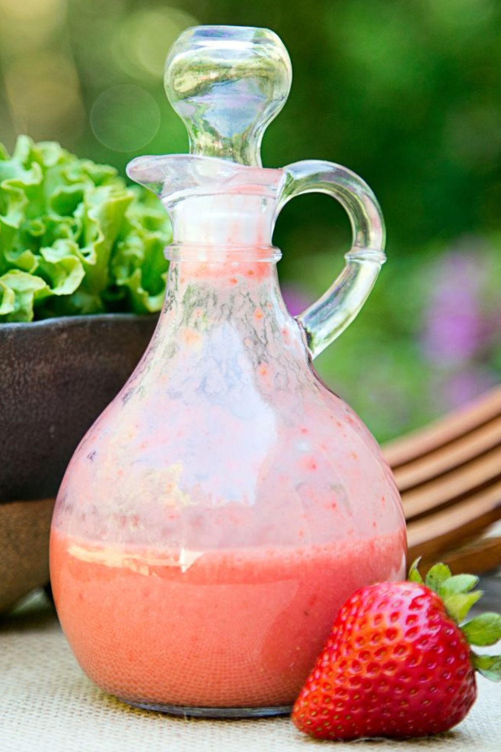 Erdbeer-Basilikum-Dressing Glaskaraffe mit rosa Dressing, daneben eine Erdbeere. Im Hintergrund ein Salatblatt in einer Schüssel.