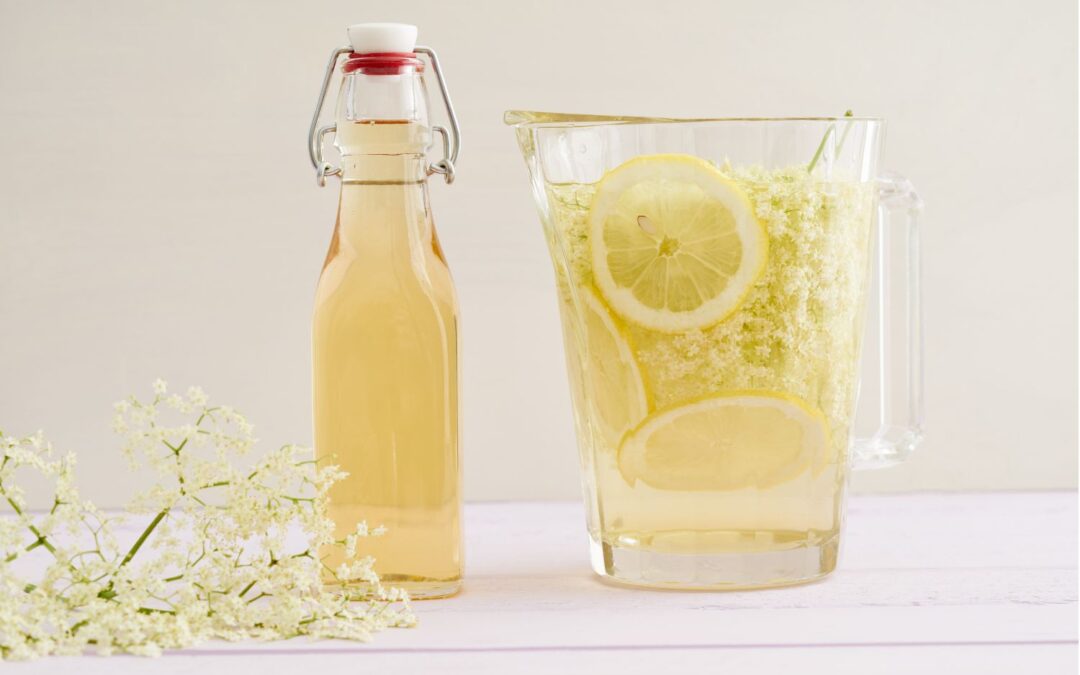 Karaffe und Flasche mit Holunderblütensirup, Zitronenscheiben und Holunderblüten auf einem hellen Tisch.