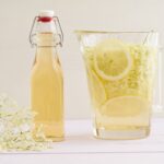 Karaffe und Flasche mit Holunderblütensirup, Zitronenscheiben und Holunderblüten auf einem hellen Tisch.
