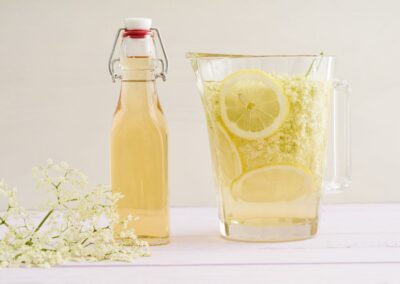 Karaffe und Flasche mit Holunderblütensirup, Zitronenscheiben und Holunderblüten auf einem hellen Tisch.