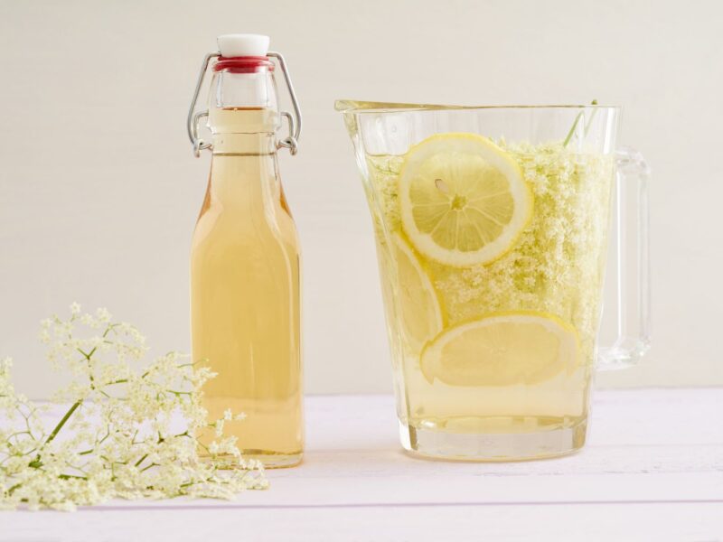 Karaffe und Flasche mit Holunderblütensirup, Zitronenscheiben und Holunderblüten auf einem hellen Tisch.
