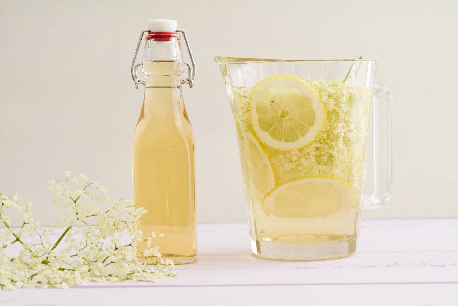 Karaffe und Flasche mit Holunderblütensirup, Zitronenscheiben und Holunderblüten auf einem hellen Tisch.