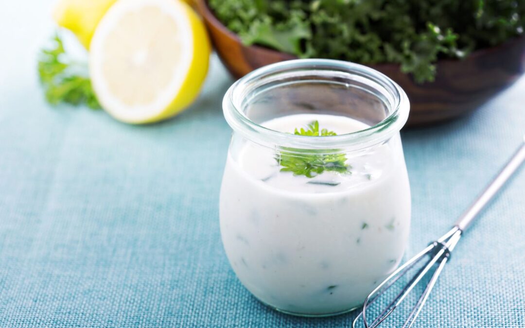 Ein Glas Joghurt-Dressing mit Kräutern auf blauem Tuch, daneben eine halbe Zitrone und eine Schüssel mit grünem Salat.