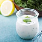Ein Glas Joghurt-Dressing mit Kräutern auf blauem Tuch, daneben eine halbe Zitrone und eine Schüssel mit grünem Salat.