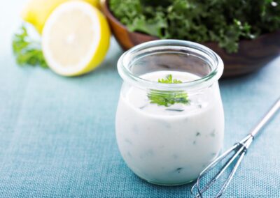 Ein Glas Joghurt-Dressing mit Kräutern auf blauem Tuch, daneben eine halbe Zitrone und eine Schüssel mit grünem Salat.