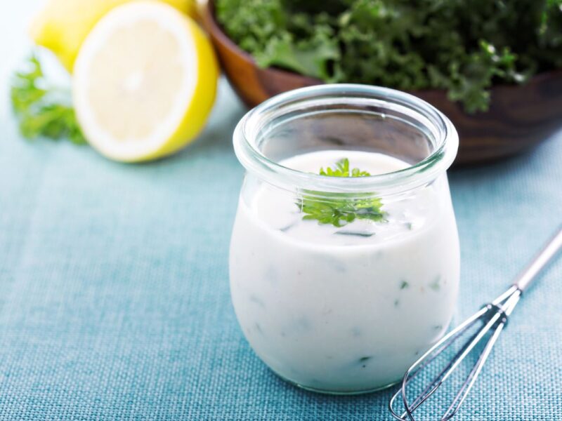 Ein Glas Joghurt-Dressing mit Kräutern auf blauem Tuch, daneben eine halbe Zitrone und eine Schüssel mit grünem Salat.