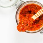 Geöffnetes Glas mit roter Sauce, Holzlöffel darin. Draufsicht, weiße Hintergrund.