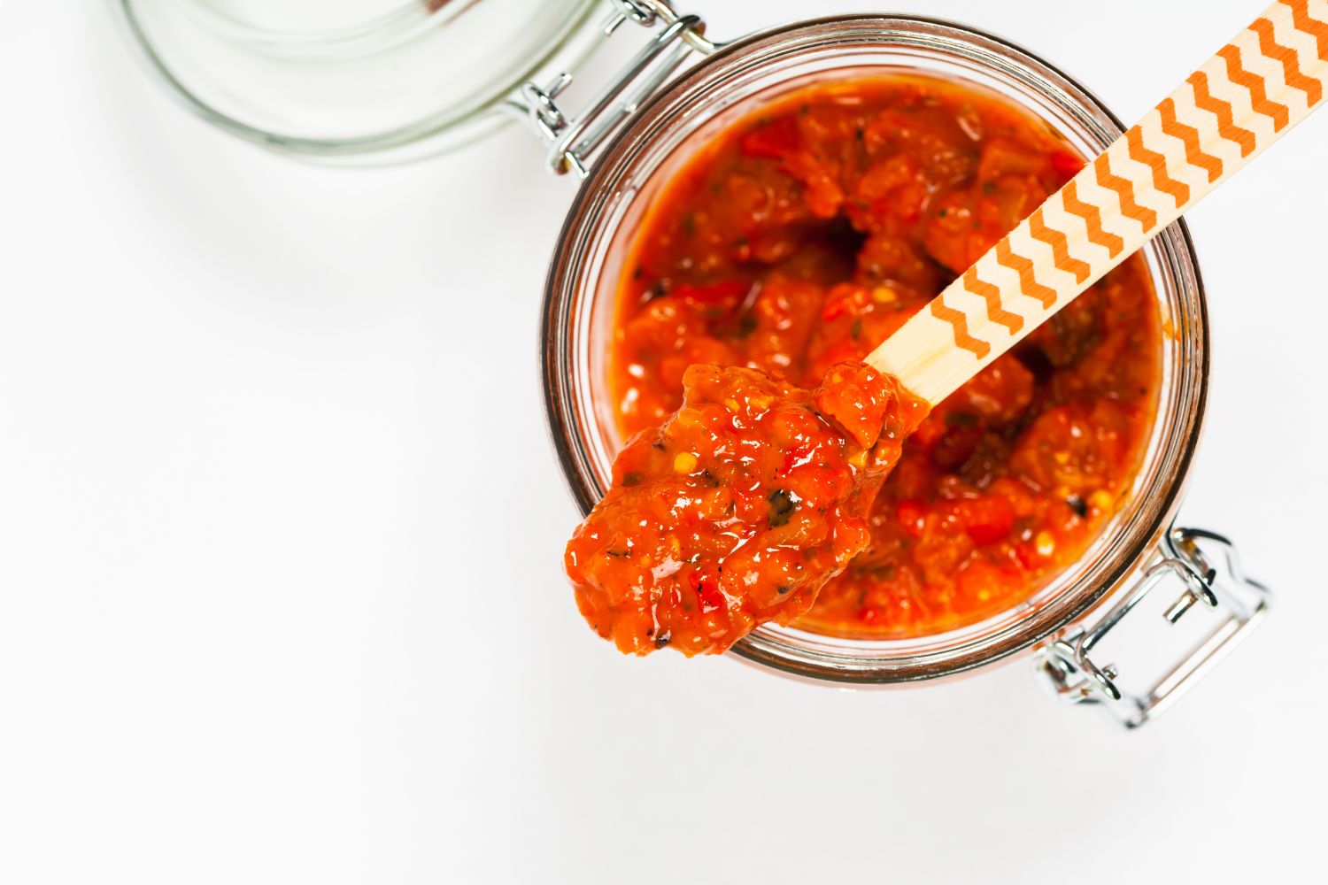 Aufstrich mit Paprika und frischen Kräutern Geöffnetes Glas mit roter Sauce, Holzlöffel darin. Draufsicht, weiße Hintergrund.