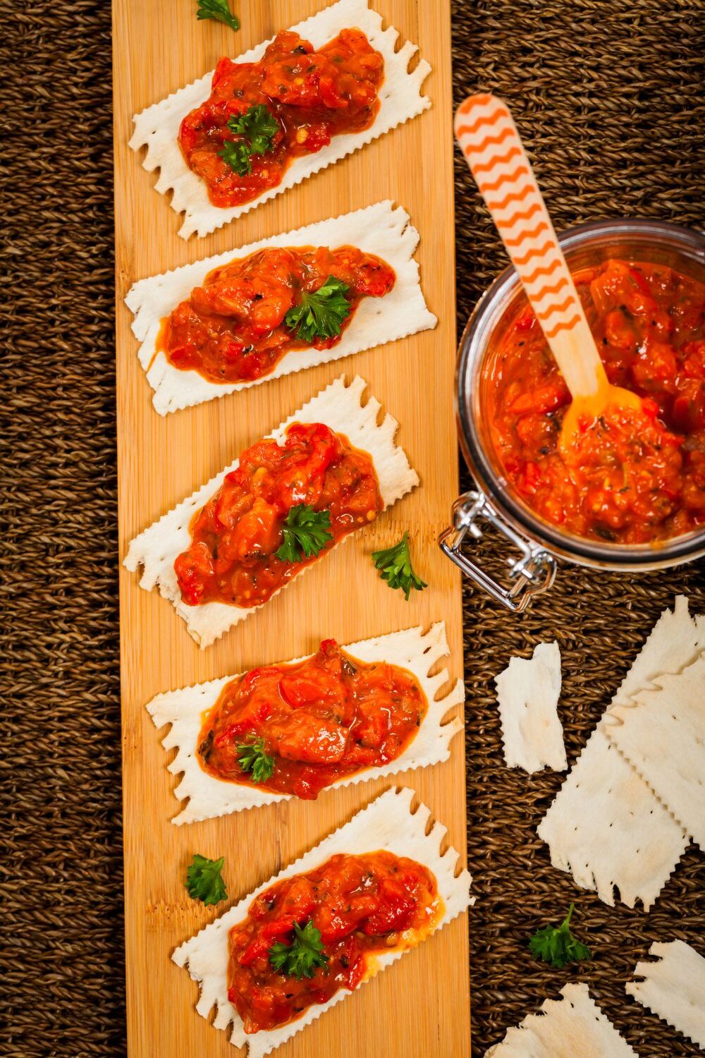 Aufstrich mit Paprika und frischen Kräutern Knusprige Cracker mit roter Tomaten-Paprika-Sauce auf einem Holzbrett. Daneben ein Glas mit der Sauce und Löffel.